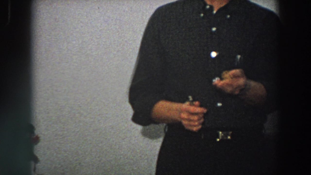 The man holds a small light-colored object closer to his body in a grainy 8mm domestic interior shot.