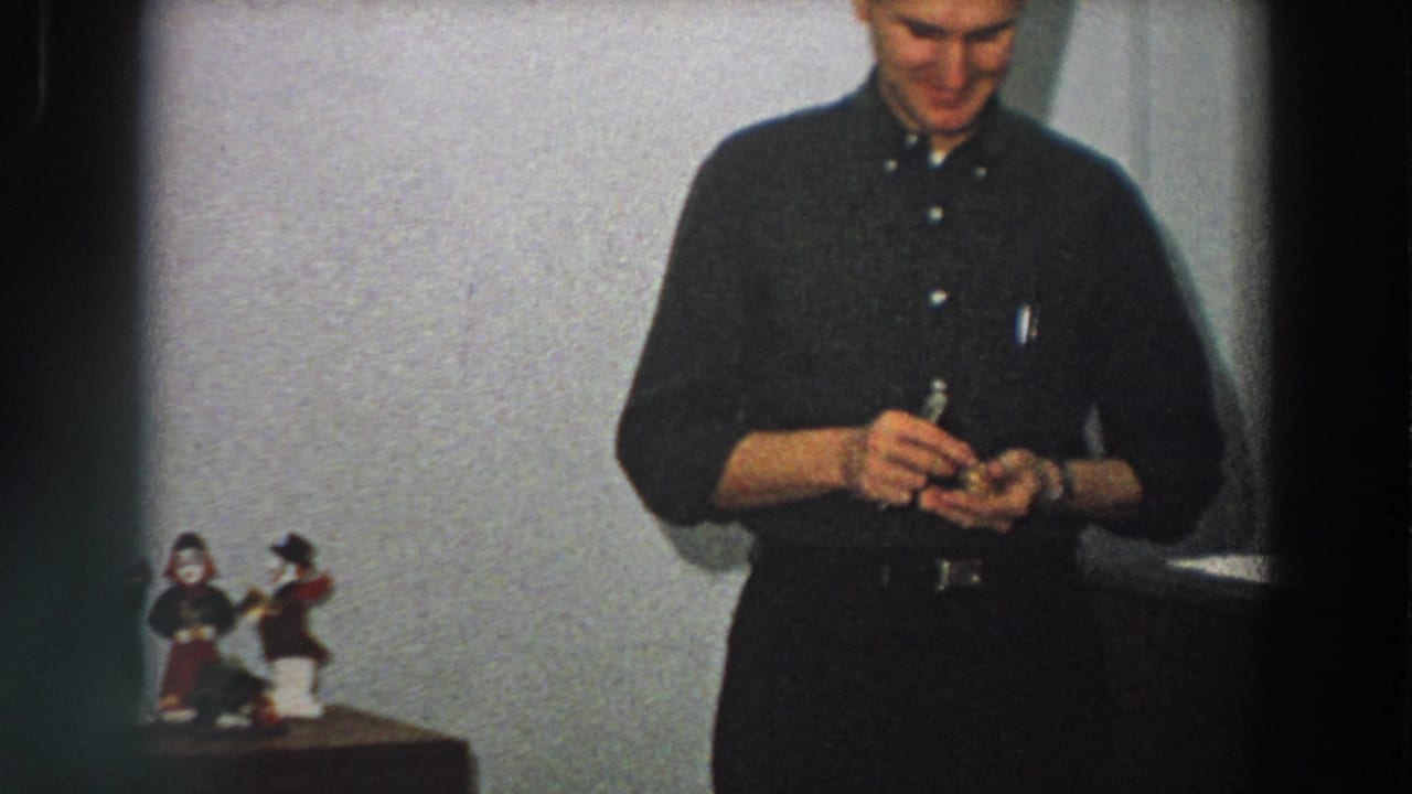 Man in dark clerical-style clothing handles a small object near tabletop religious figurines.