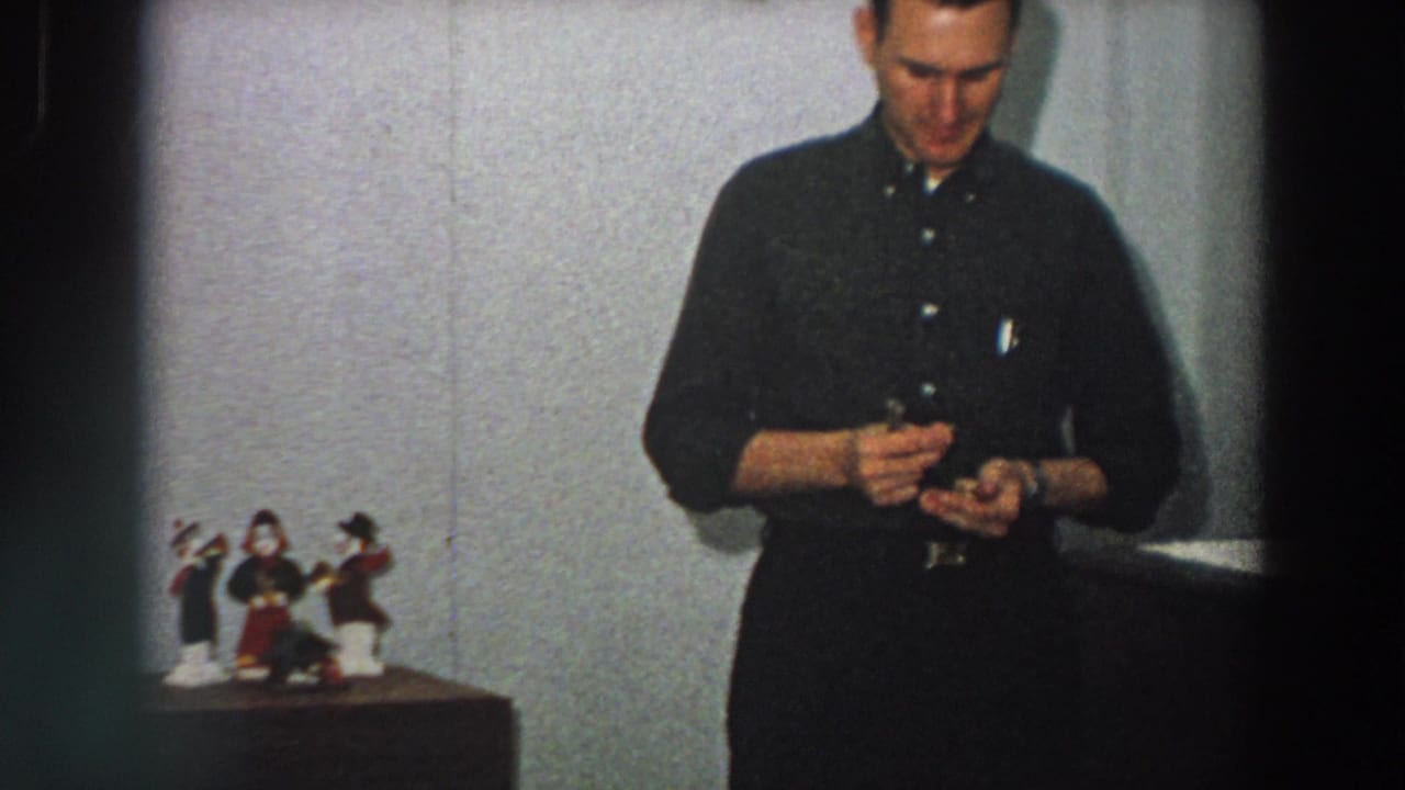 The man remains near the wall and tabletop figures, focused on a small object, in color 8mm home-movie footage identified as Italy, 1958.