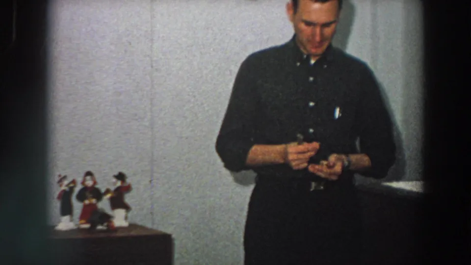 The man remains near the wall and tabletop figures, focused on a small object, in color 8mm home-movie footage identified as Italy, 1958.