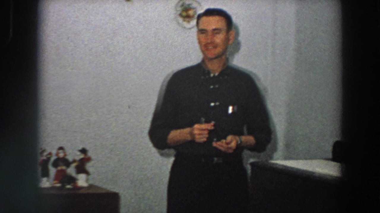 A man in dark clerical-style clothing smiles while holding a small object, with religious figurines visible on a nearby tabletop in a private interior.