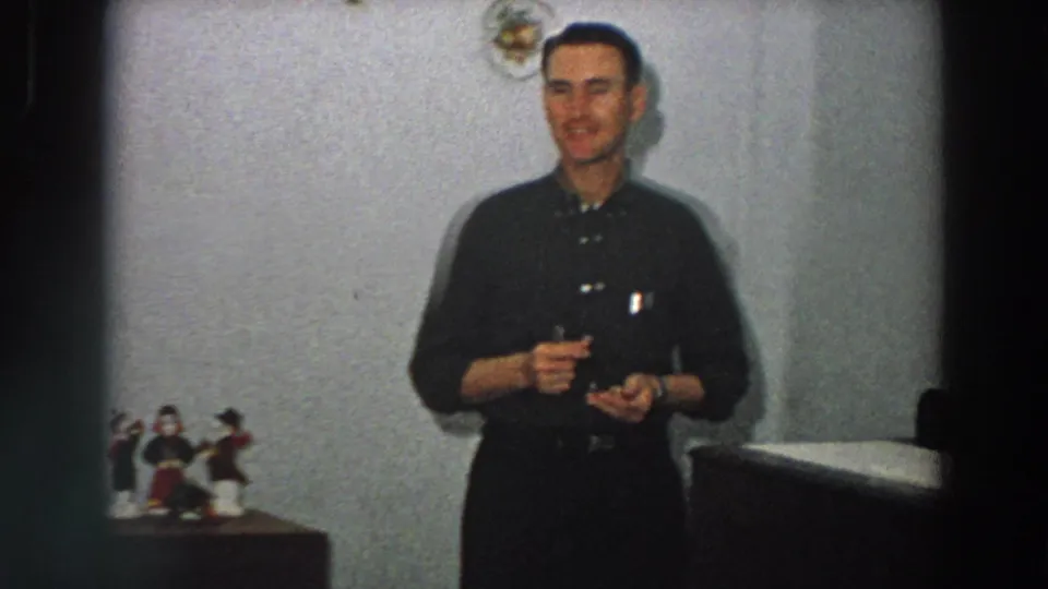 A man in dark clerical-style clothing smiles while holding a small object, with religious figurines visible on a nearby tabletop in a private interior.