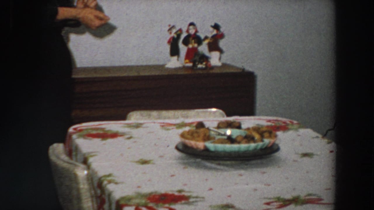 1958 Italy 8mm frame showing a floral dining table, food dish, wooden sideboard, figurines, and a partial person entering at left.