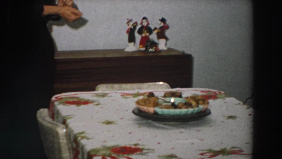1958 Italy 8mm frame showing a floral dining table, food dish, wooden sideboard, figurines, and a partial person entering at left.
