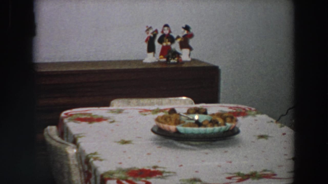 1958 Italy home-movie view of a floral-covered dining table, dish of food, brown sideboard, and small figurines against a plain wall.