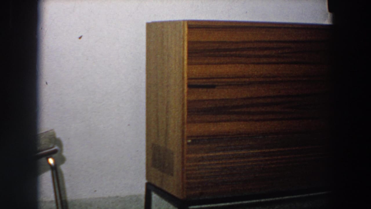 1958 Italy 8mm frame focused on a brown wooden sideboard with striped grain, plain wall, and a small table edge in the foreground.