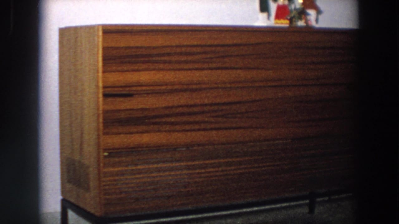 1958 Italy home-movie frame showing the brown sideboard more fully, with figurines partly cropped at the top and dark film edges.