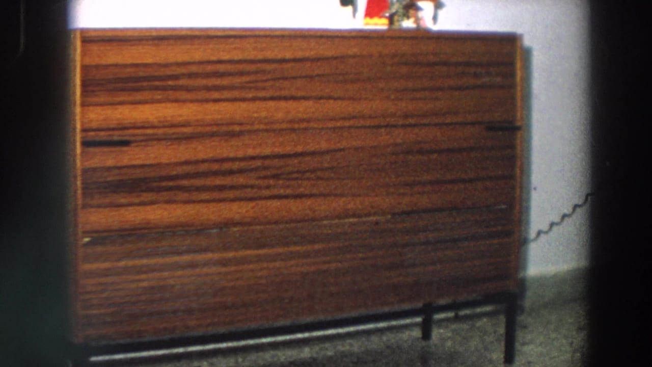 1958 Italian home-movie frame showing a brown striped wooden console table against a pale interior wall.