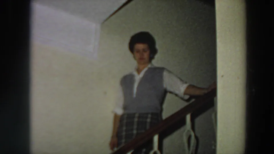 Adult woman in a grey vest, white blouse, and plaid skirt stands or descends on an indoor staircase in Italy, 1958.