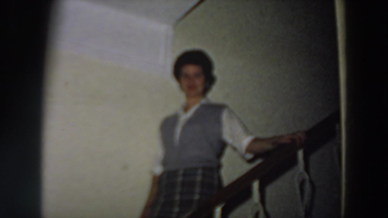 In 1958 Italy home-movie footage, a woman in a blouse, vest, and plaid skirt stands near a stair railing in soft focus.