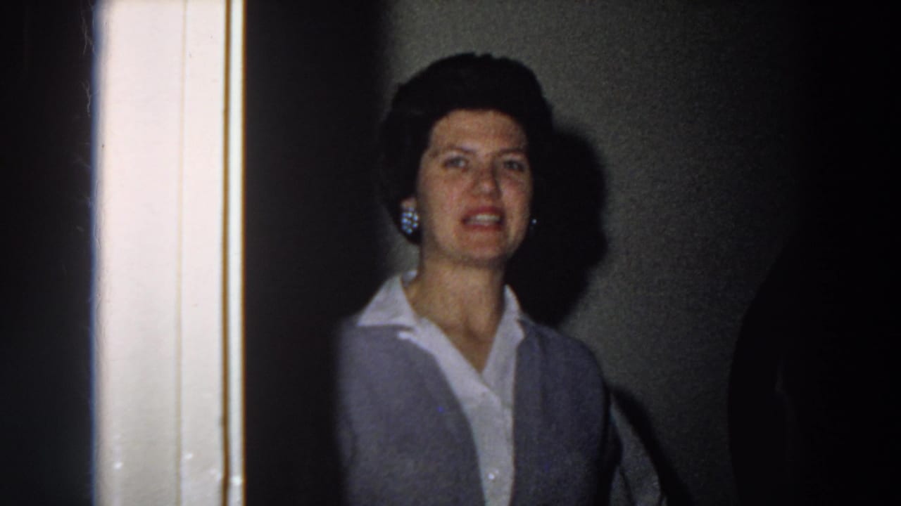 Closer 1958 Italy home-movie frame of a woman near the stairwell, with earrings, dark styled hair, blouse, and vest visible.