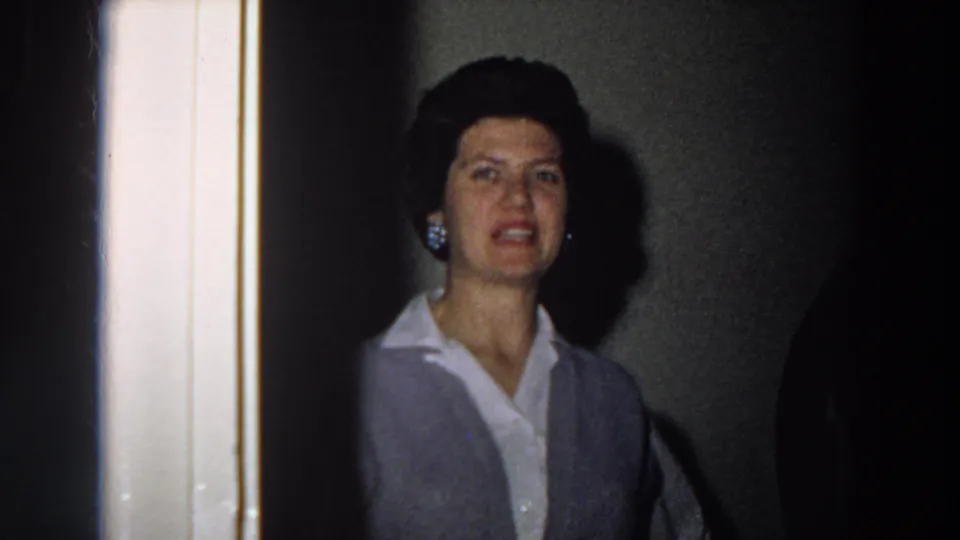 Closer 1958 Italy home-movie frame of a woman near the stairwell, with earrings, dark styled hair, blouse, and vest visible.
