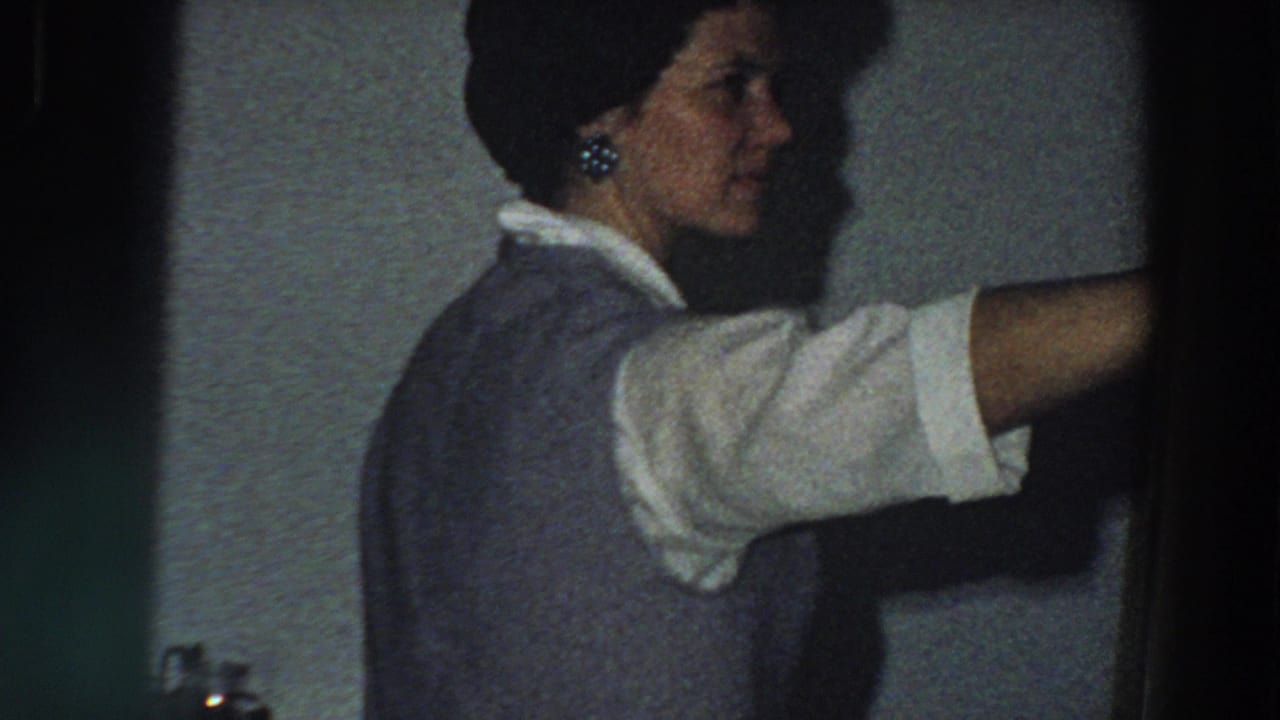 In a 1958 Italian home interior, a woman in profile reaches off-frame beside a plain wall, filmed in grainy 8mm.