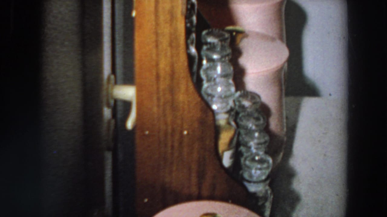 The camera pans along the wooden shelf, showing glass jars and a pink household container in a 1958 Italian interior.