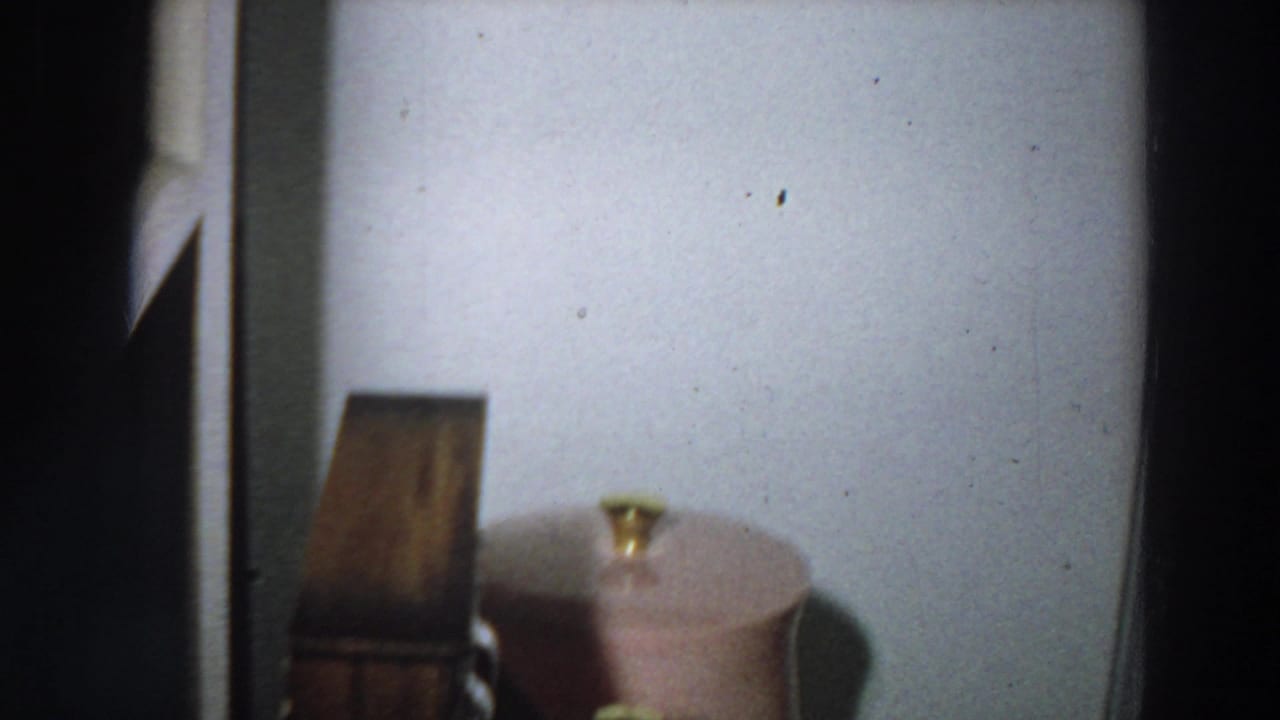 A low, slightly blurred 8mm view shows a pink lidded container and small wooden object against a plain wall in the home.