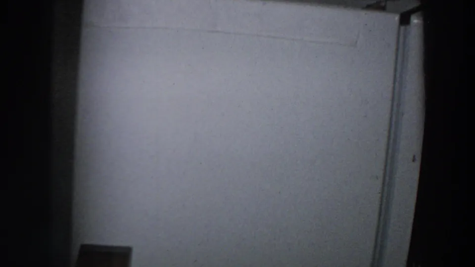 1958 8mm frame from Italy showing a dim kitchen corner with a white refrigerator door and shadowed framing.
