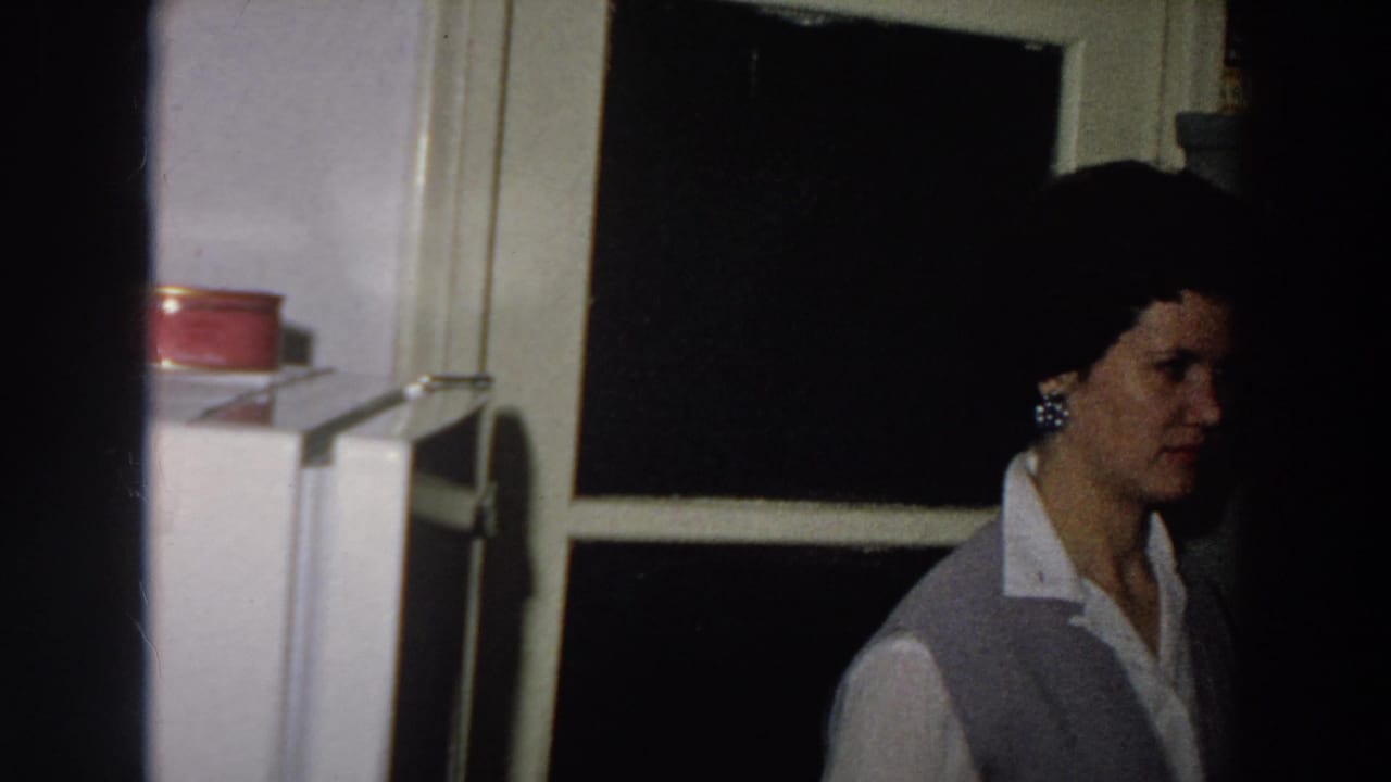 1958 8mm frame from Italy showing a woman in a light blouse and grey vest standing near a refrigerator and dark window.