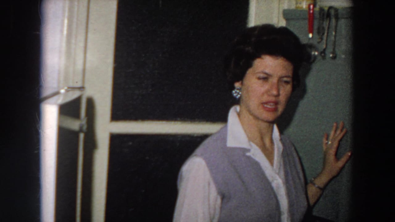1958 8mm frame from Italy showing a woman in a grey vest near a dark window and wall-mounted kitchen utensils.