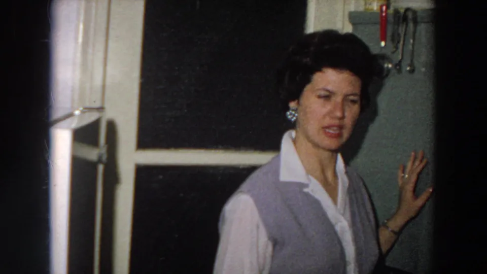 1958 8mm frame from Italy showing a woman in a grey vest near a dark window and wall-mounted kitchen utensils.