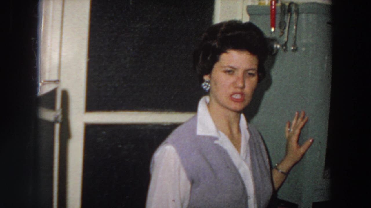 In a 1958 Italy home-movie frame, a woman in a white collared shirt and lavender cardigan gestures beside a green kitchen hutch.