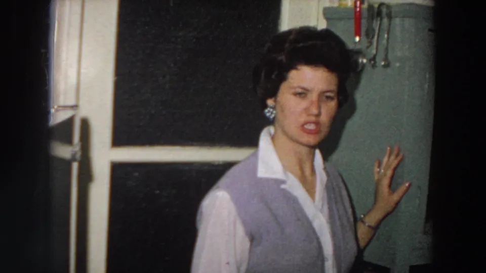 In a 1958 Italy home-movie frame, a woman in a white collared shirt and lavender cardigan gestures beside a green kitchen hutch.