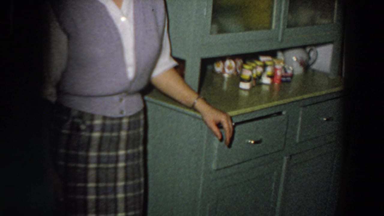 The camera crops the woman as she stands near green cabinetry, drawers, glass doors, and packaged kitchen goods in the 1958 Italy interior.