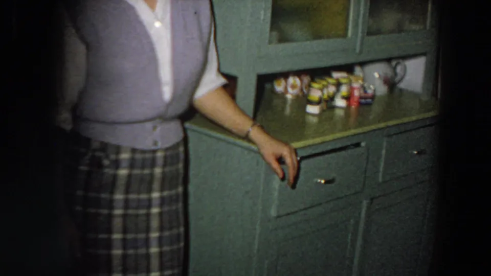 The camera crops the woman as she stands near green cabinetry, drawers, glass doors, and packaged kitchen goods in the 1958 Italy interior.