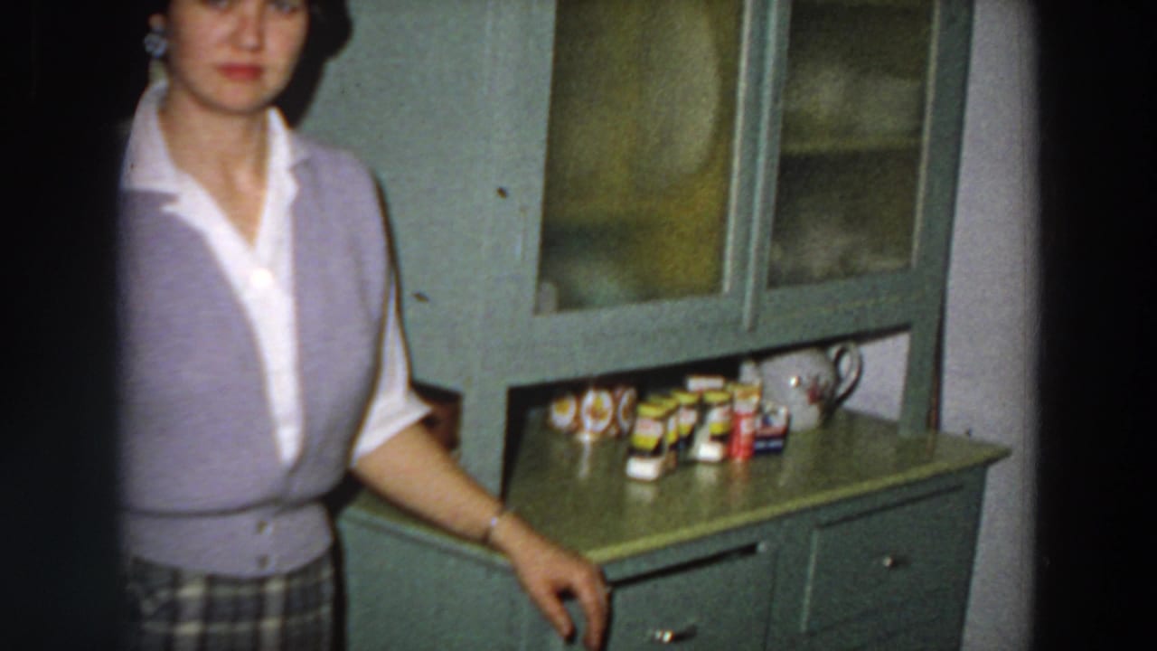 A cropped home-movie view shows the woman beside a green cabinet topped with small tins and containers in the Italy kitchen setting.