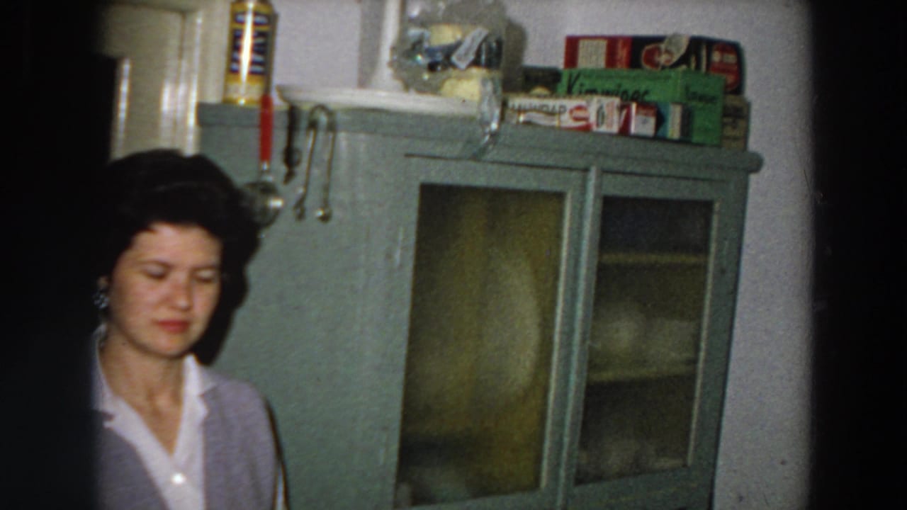 The woman appears lower in the frame beside a tall green glass-front kitchen cabinet, with hanging utensils and packaged goods above.
