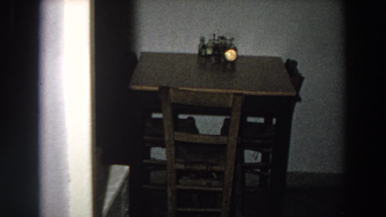 1958 Italian home-movie frame showing a small brown table, wooden chairs, and a glowing lamp in a modest interior.