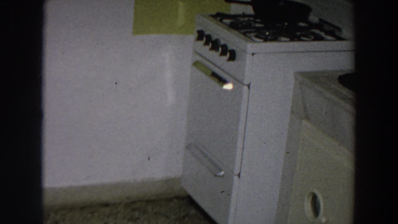 1958 Italian home-movie frame showing a white stove, black cookware, green wall panel, and adjacent counter in a private kitchen.