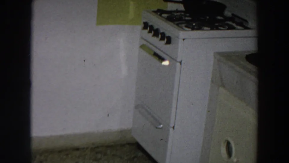 1958 Italian home-movie frame showing a white stove, black cookware, green wall panel, and adjacent counter in a private kitchen.