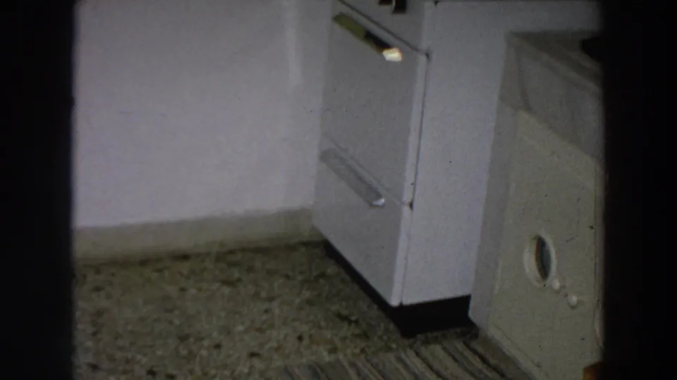 1958 Italy home-movie frame showing a white stove, speckled floor, striped rug, and dark doorway edge in a private kitchen.