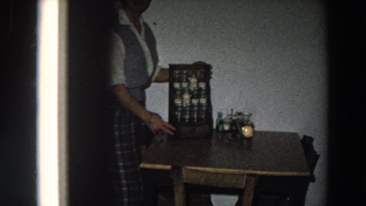 1958 Italy kitchen view with a woman beside a wooden table, small spice cabinet, and glowing candle or lamp.