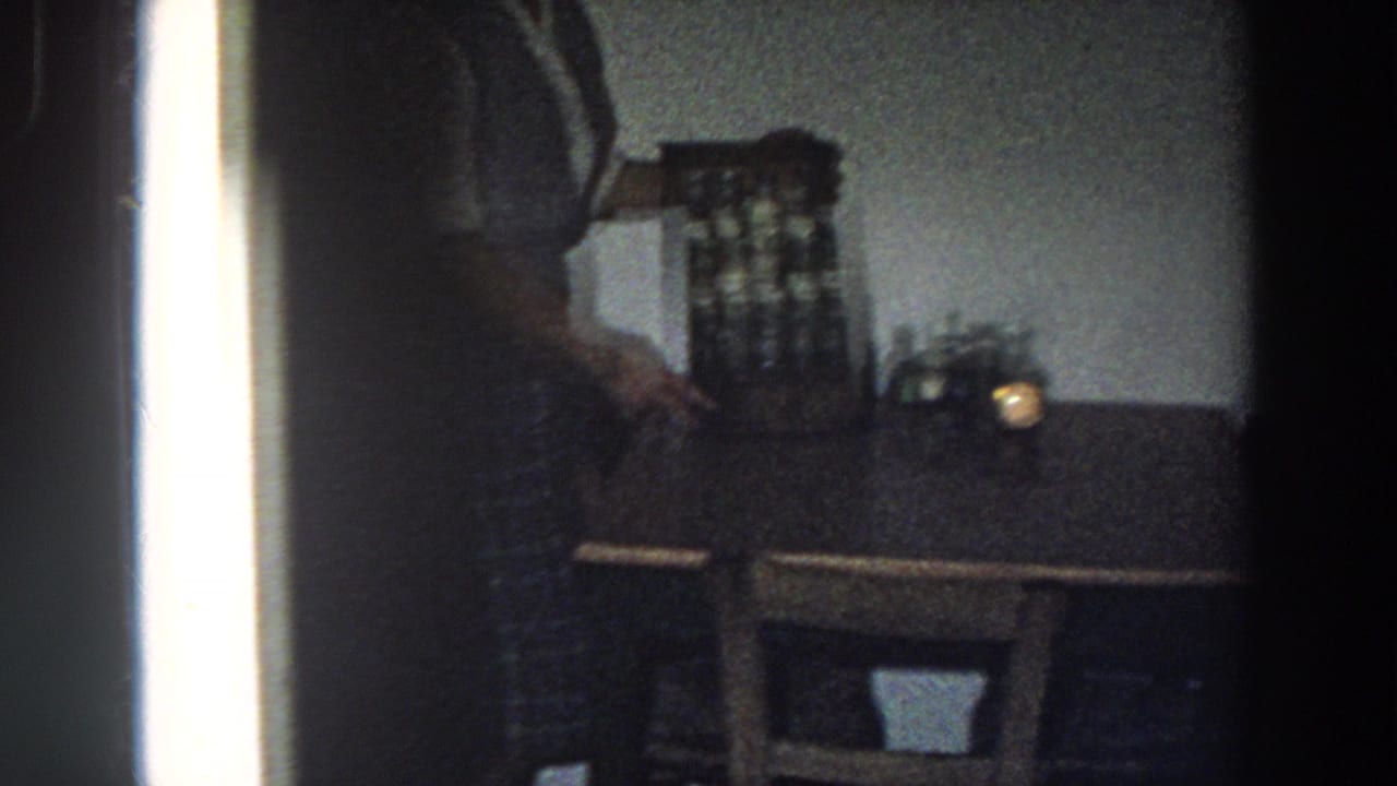 Dim 1958 Italy home-movie frame of a woman reaching near a spice cabinet on a wooden table in a private kitchen.