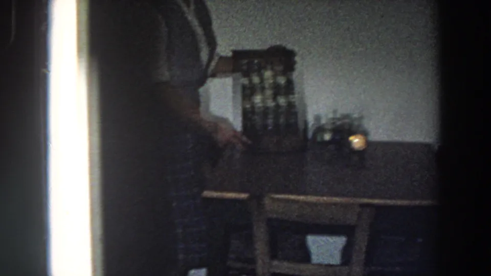 Dim 1958 Italy home-movie frame of a woman reaching near a spice cabinet on a wooden table in a private kitchen.