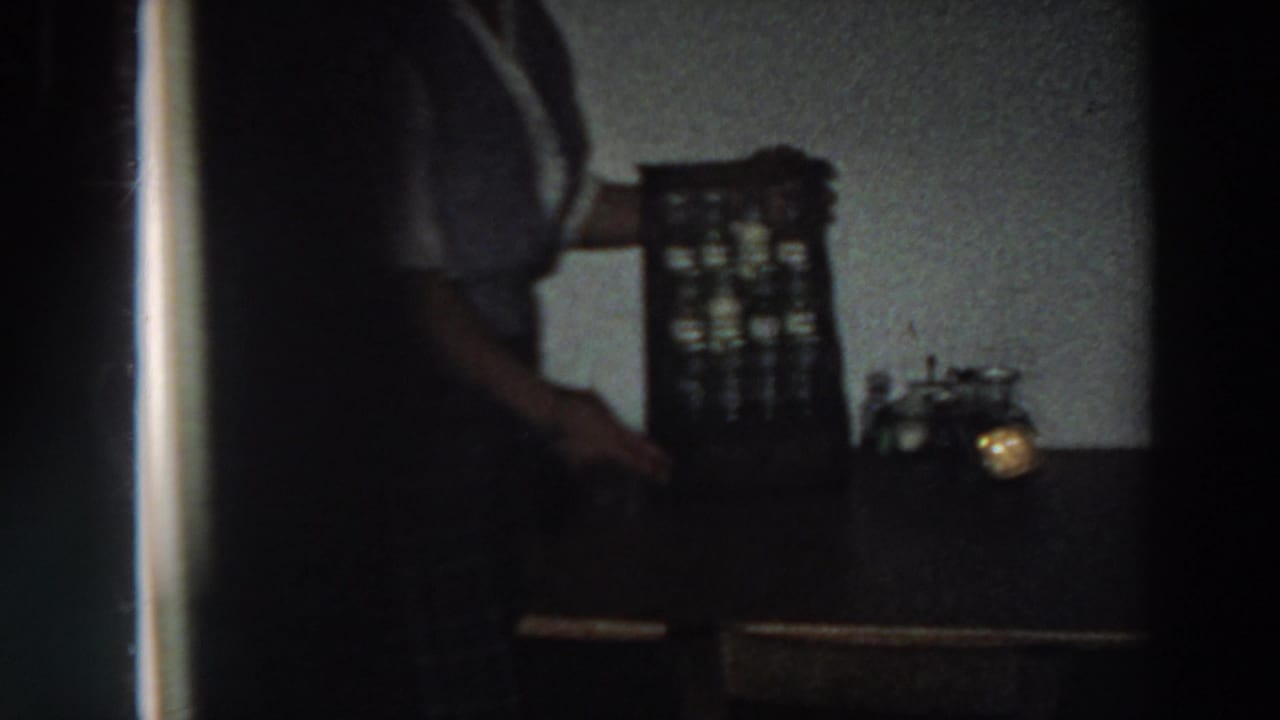 1958 Italy kitchen close view with a partially visible woman, spice cabinet, tabletop, and small glowing light in soft 8mm exposure.