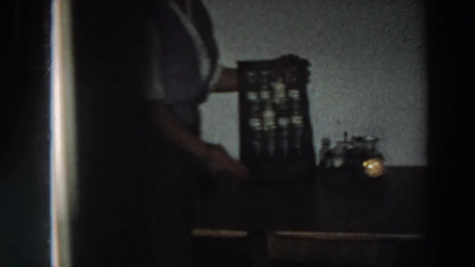 1958 Italy kitchen close view with a partially visible woman, spice cabinet, tabletop, and small glowing light in soft 8mm exposure.