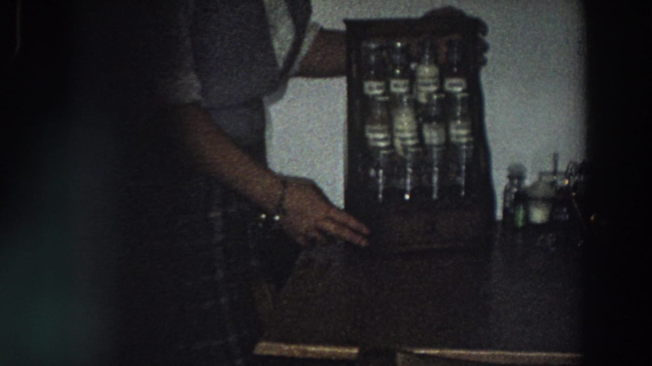 In a dim Italy 1958 home-movie frame, a woman stands near a tabletop wooden holder containing labeled glass bottles.