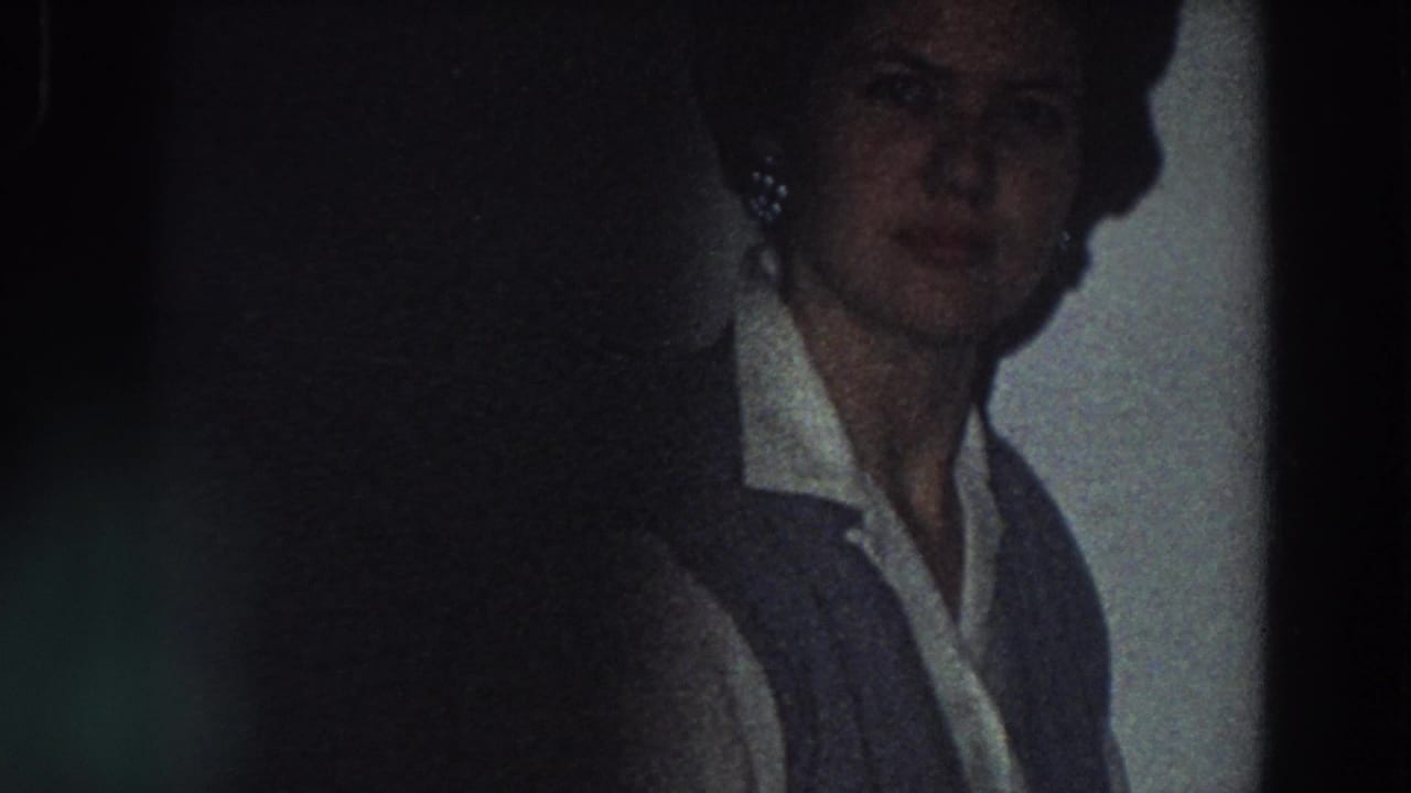 In 1958 Italy home-movie footage, a woman stands near a tiled kitchen wall in a white collared shirt and dark vest.