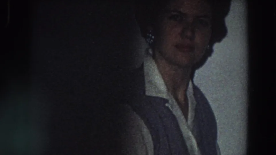 In 1958 Italy home-movie footage, a woman stands near a tiled kitchen wall in a white collared shirt and dark vest.
