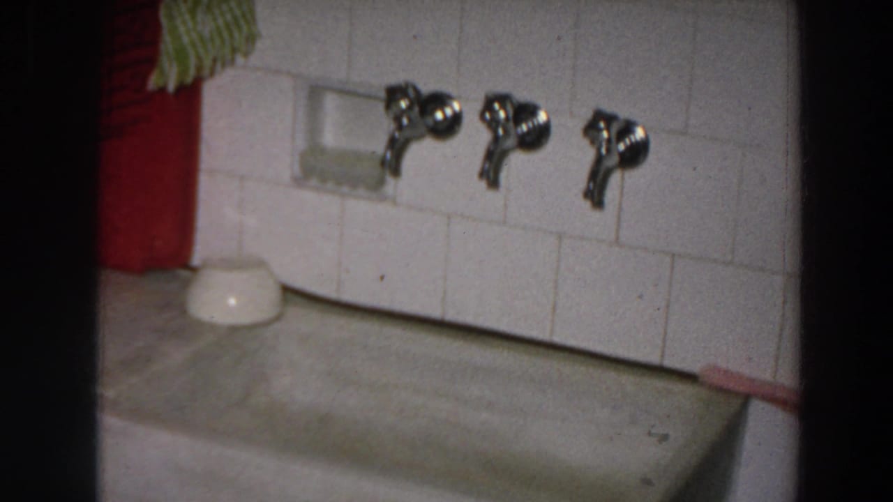 A tiled kitchen wall with three chrome faucets sits above a deep sink basin, with cloths and a small white bowl visible.