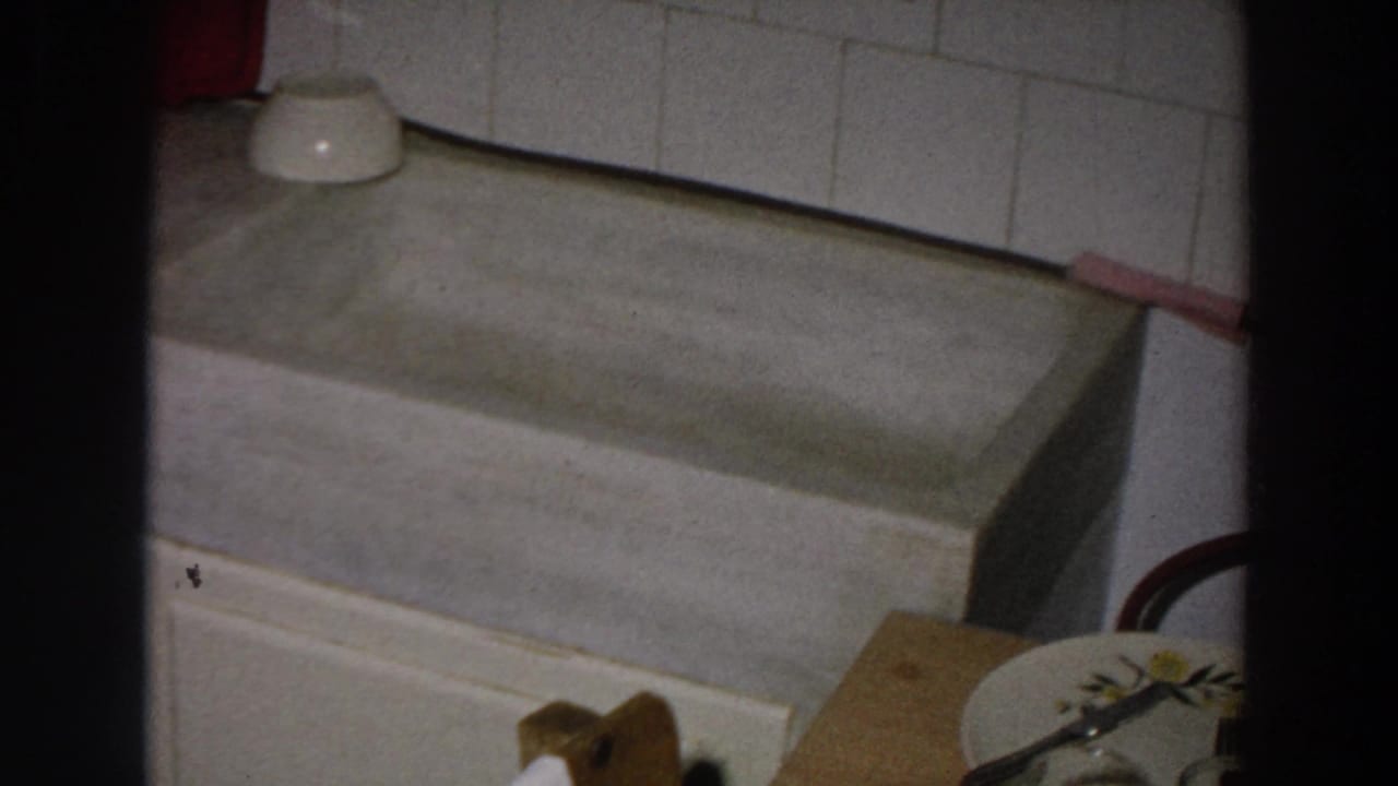 The camera settles on the deep sink basin and tiled wall, with a small bowl, cloth, and partial dish visible at frame edge.