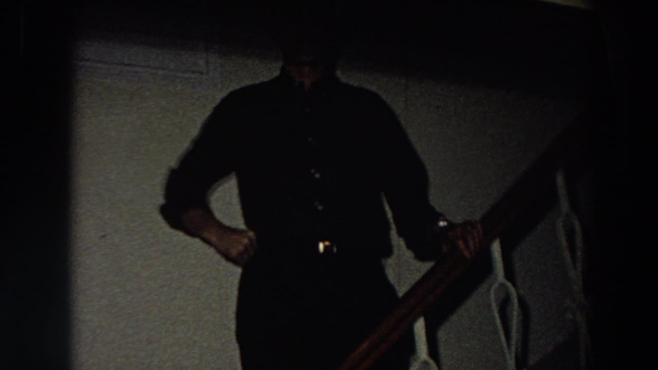 In a dim 1958 Italy home-movie frame, an adult man in a dark shirt and trousers stands near a wooden stair rail.