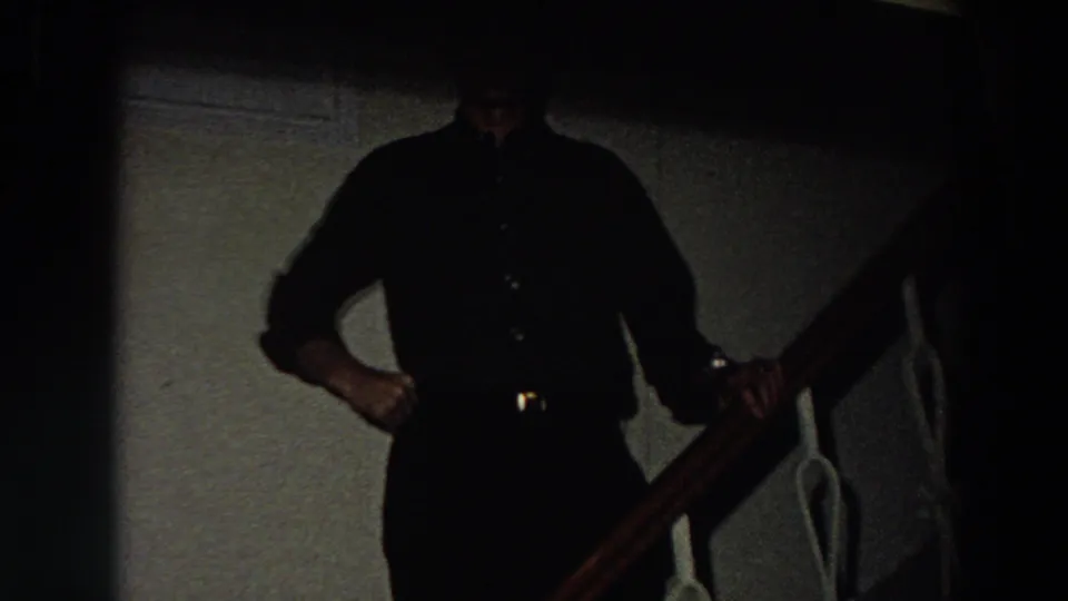 In a dim 1958 Italy home-movie frame, an adult man in a dark shirt and trousers stands near a wooden stair rail.