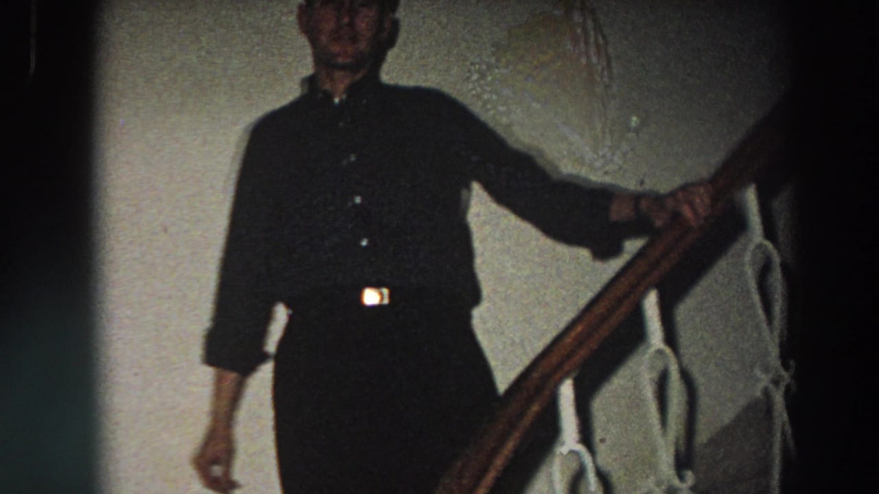 The man moves down the staircase in a dark button-up shirt, partly lit against a pale interior wall.