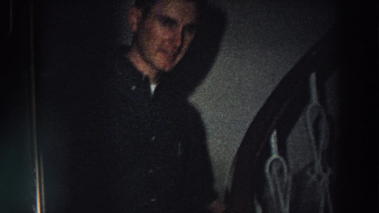 Man in a dark coat standing near an interior staircase in dim light.