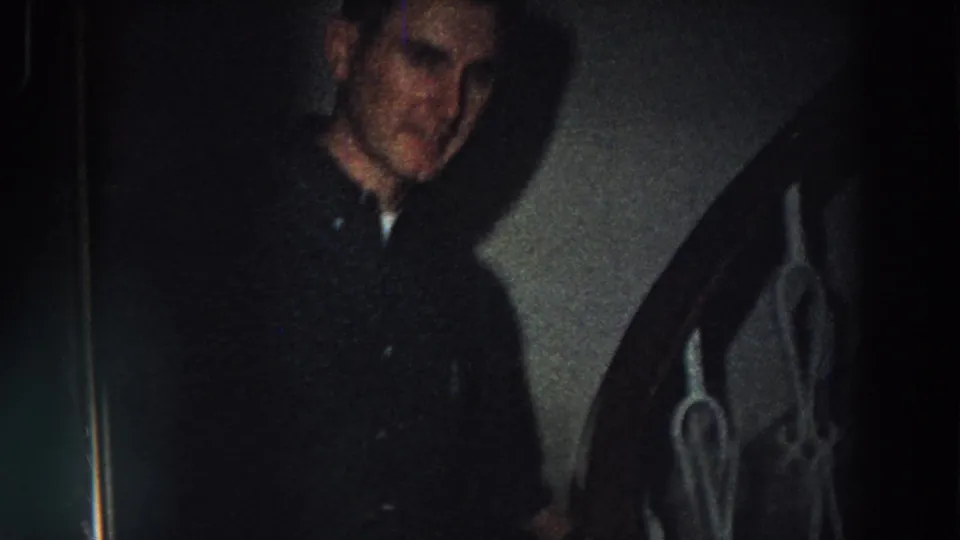 In 1958 Italy home-movie footage, a man in a dark coat stands by a staircase or metal railing inside a dim house.