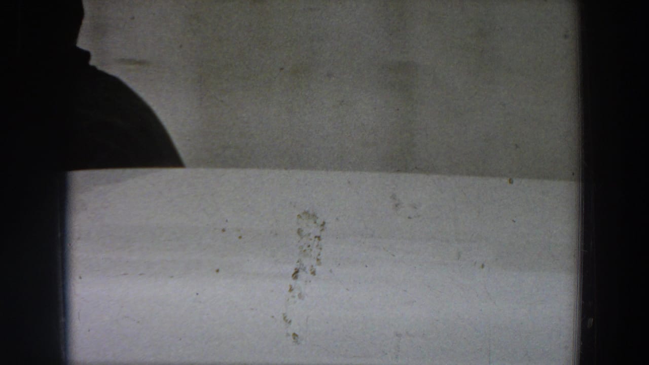 A shadowed figure or partial silhouette appears at the edge of a pale interior wall, with visible 8mm film grain and marks.