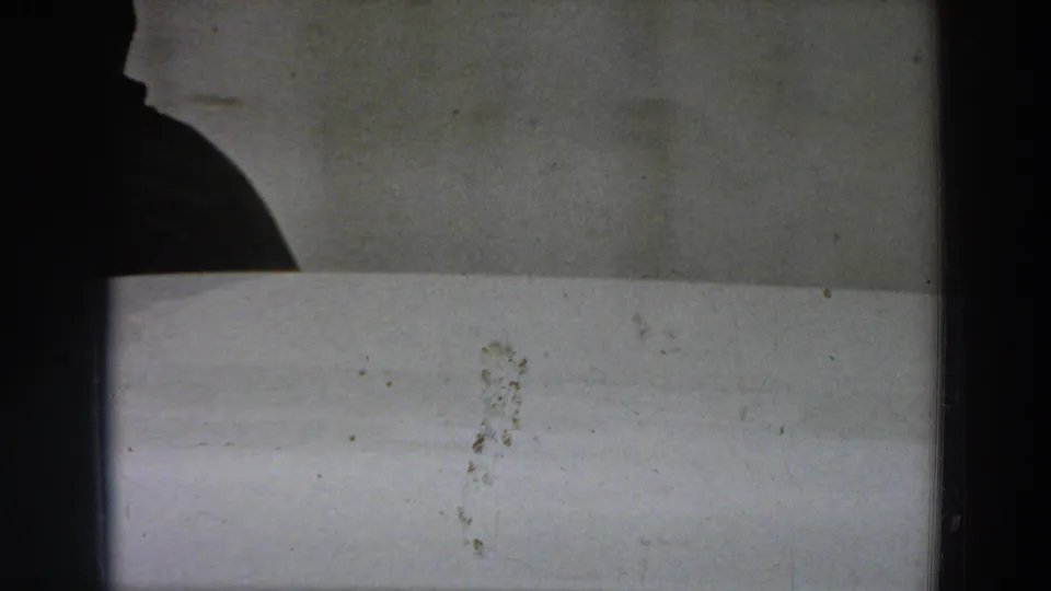 A shadowed figure or partial silhouette appears at the edge of a pale interior wall, with visible 8mm film grain and marks.